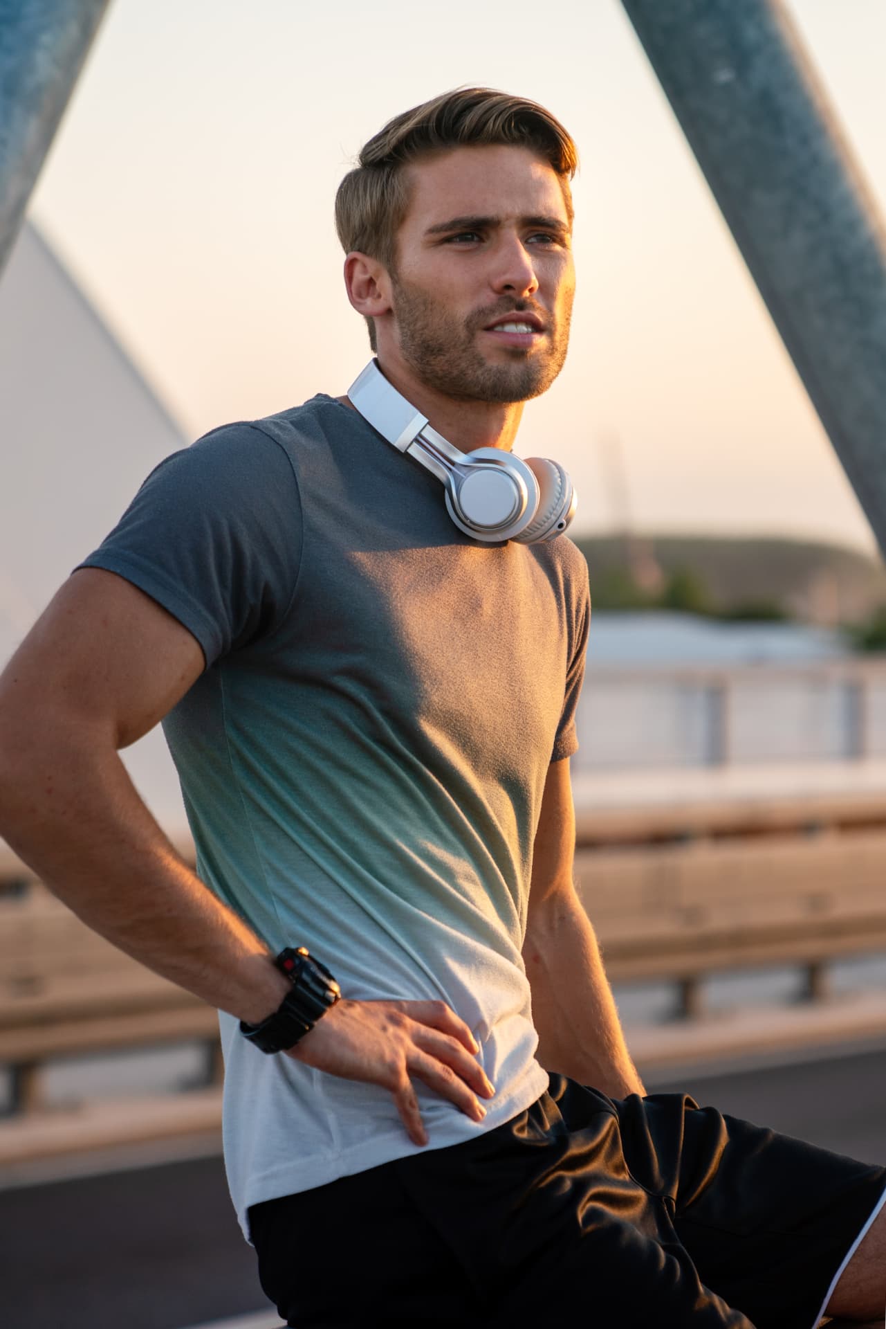 Man in a grey tee with headphones around his neck pausing outdoors at sunset